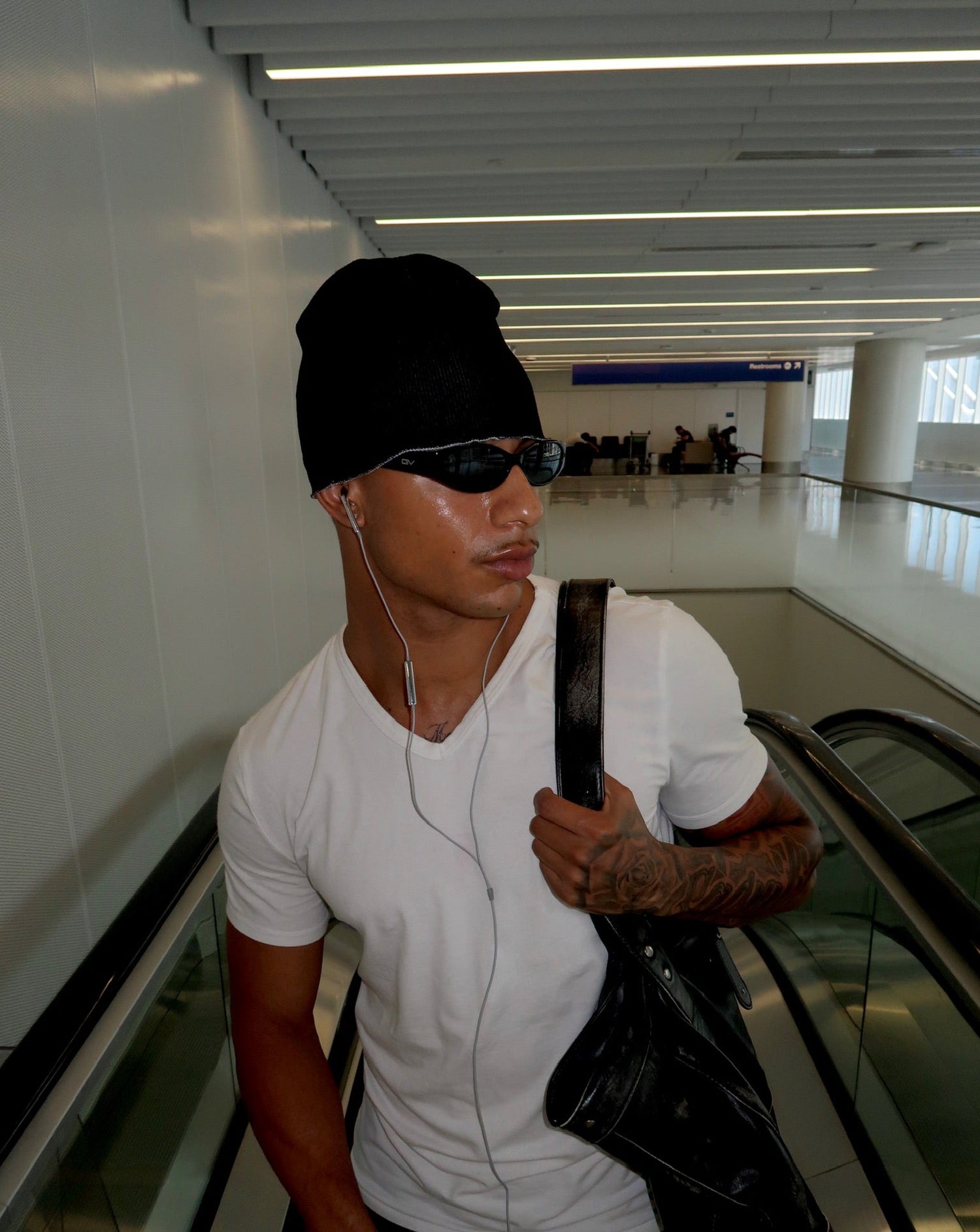 A man wearing a black beanie, black sunglasses, a white t-shirt, and carrying a black shoulder bag while riding an escalator in an airport terminal.