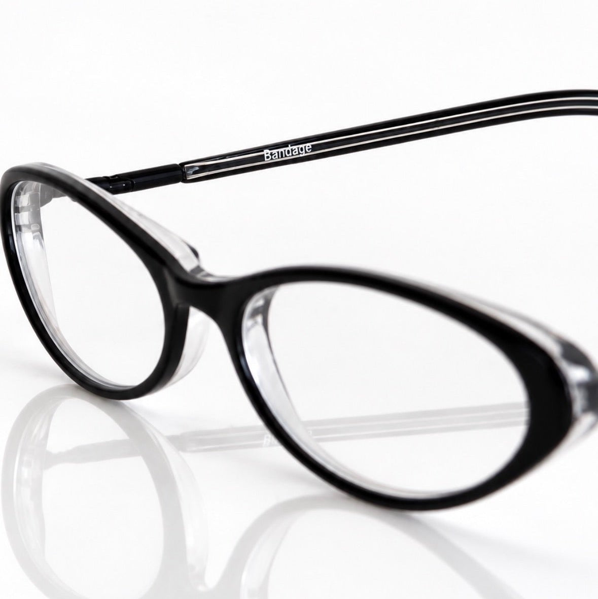 Black BANDAGE eyeglasses with clear lenses and thin silver temples, shown on a reflective white surface against a white background.