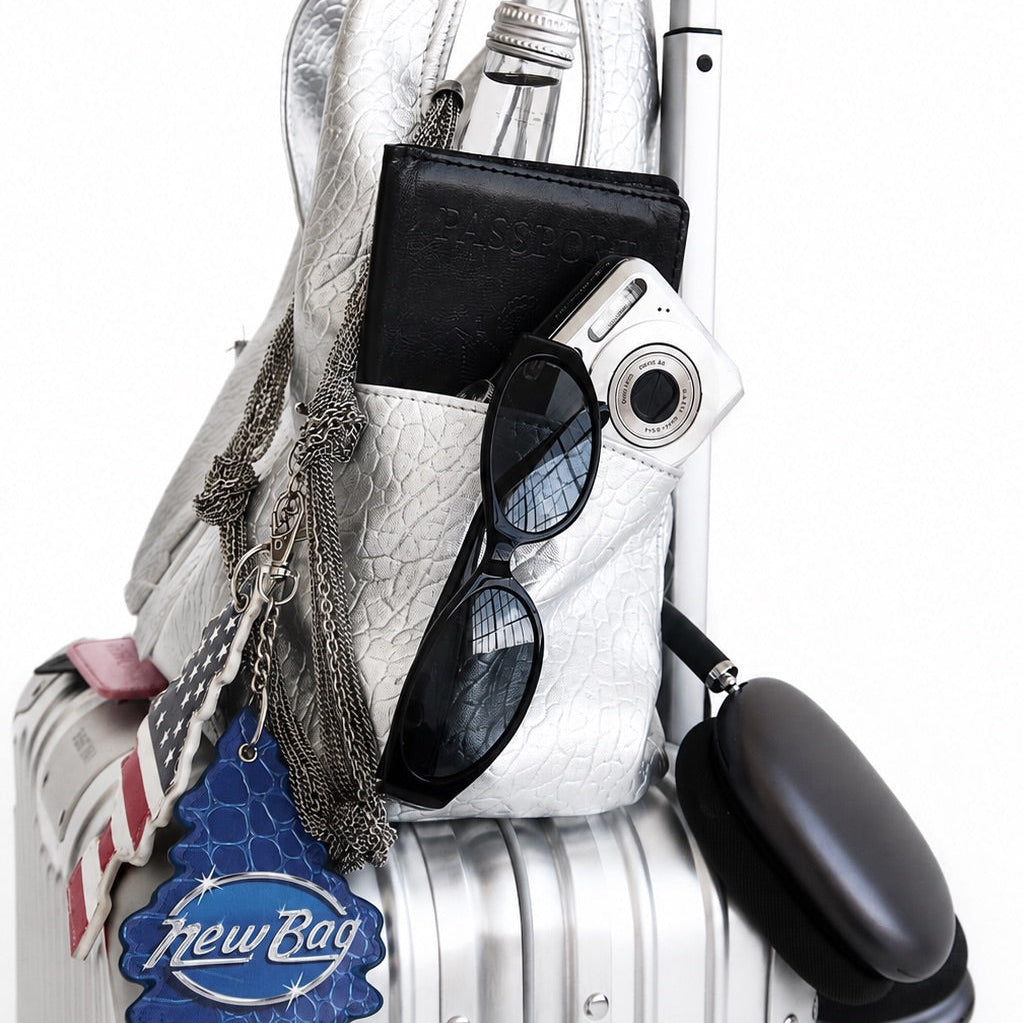 A silver textured shoulder bag packed onto a white hard-shell suitcase, alongside a black wallet, sunglasses, a silver camera, and a black pouch, with a blue 'New Bag Store' tag hanging from the bag.