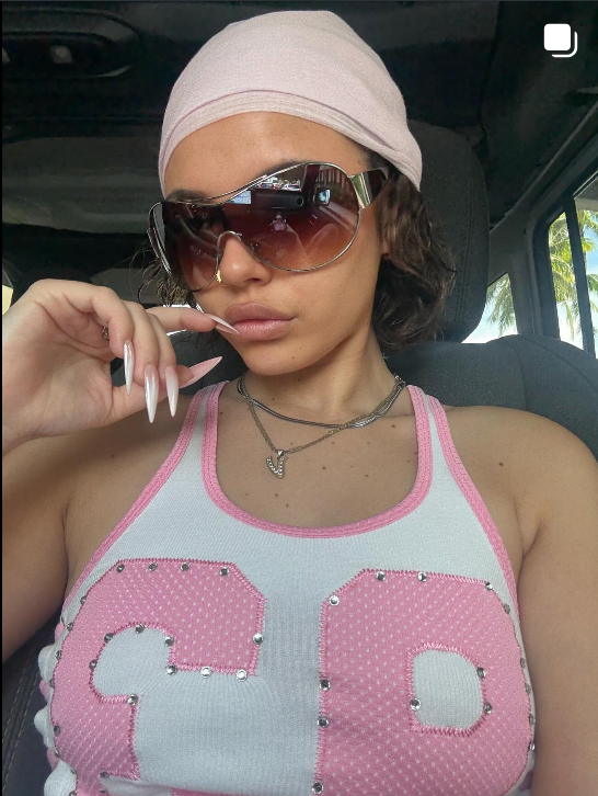 Instagram post showing Person wearing a pink and gray tank top with decorative elements, sunglasses, and a headscarf inside a vehicle.
