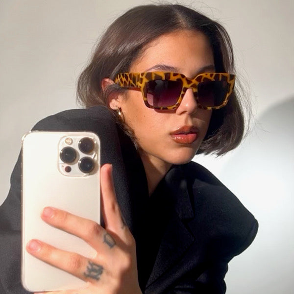 Short haired girl taking a picture on her phone wearing oversized retro SIAMESE style sunglasses from Giant Vintage 