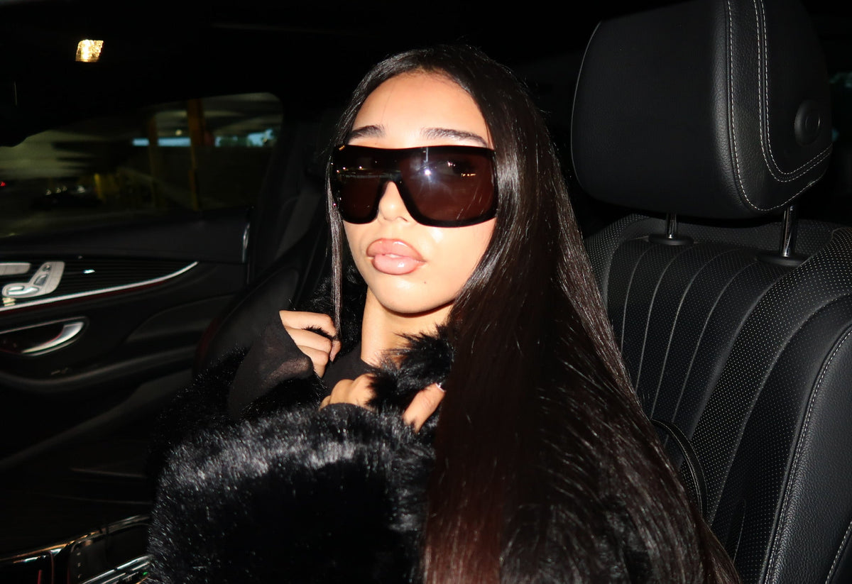 A woman with long dark hair wearing oversized dark sunglasses and a black fur-trimmed coat, seated in a black leather car interior at night.