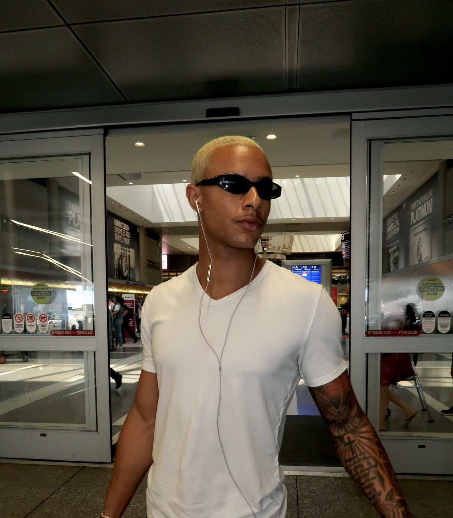 A man wearing a white crew-neck t-shirt, black sunglasses, and black jeans walks through an airport or mall entrance, holding a black bag.