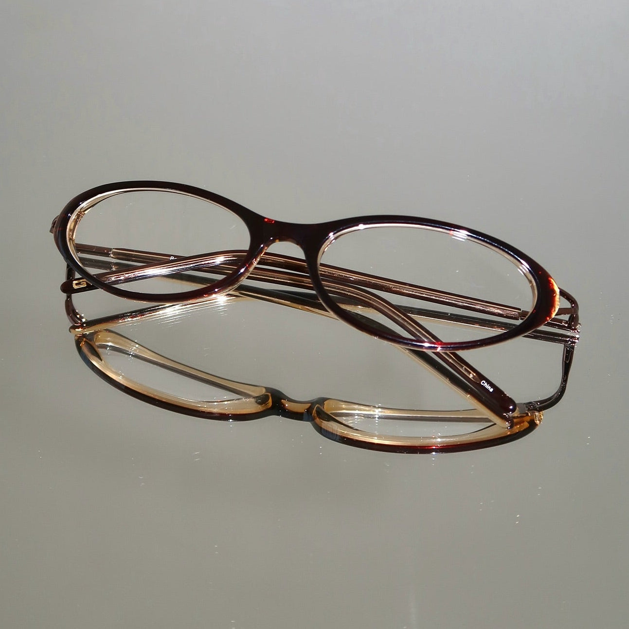 Two pairs of eyeglasses resting on a light gray reflective surface. The top pair has dark brown acetate with rose-gold-toned metal temples, and the bottom pair has tortoiseshell frames with gold-toned metal temples.