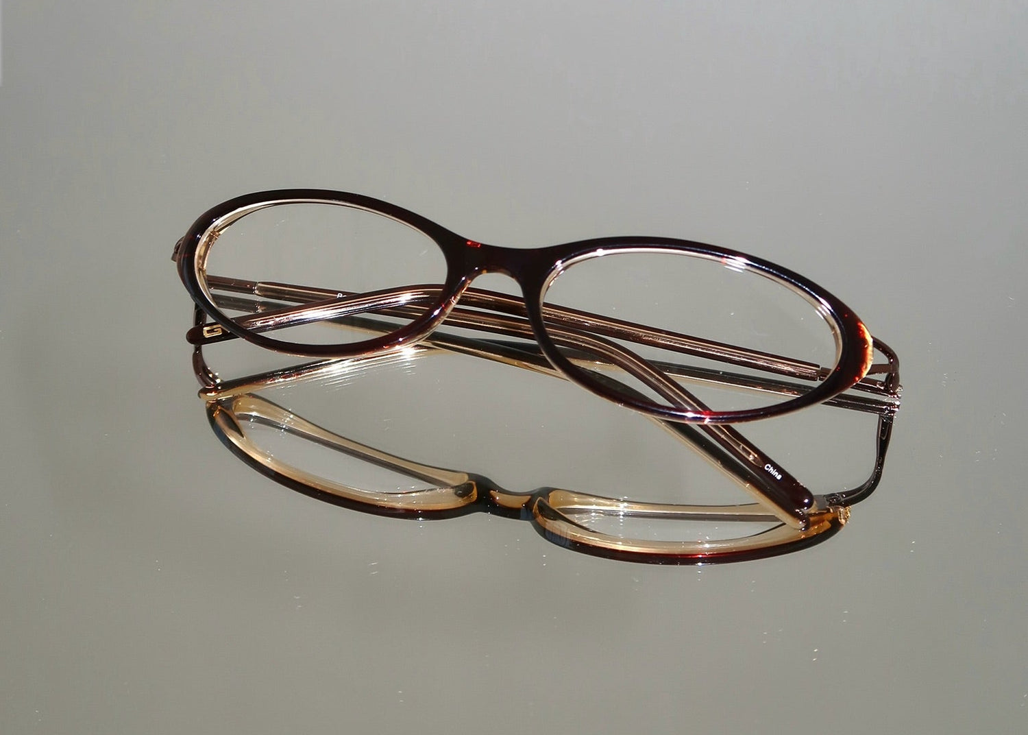 Two pairs of eyeglasses resting on a light gray reflective surface. The top pair has dark brown acetate with rose-gold-toned metal temples, and the bottom pair has tortoiseshell frames with gold-toned metal temples.