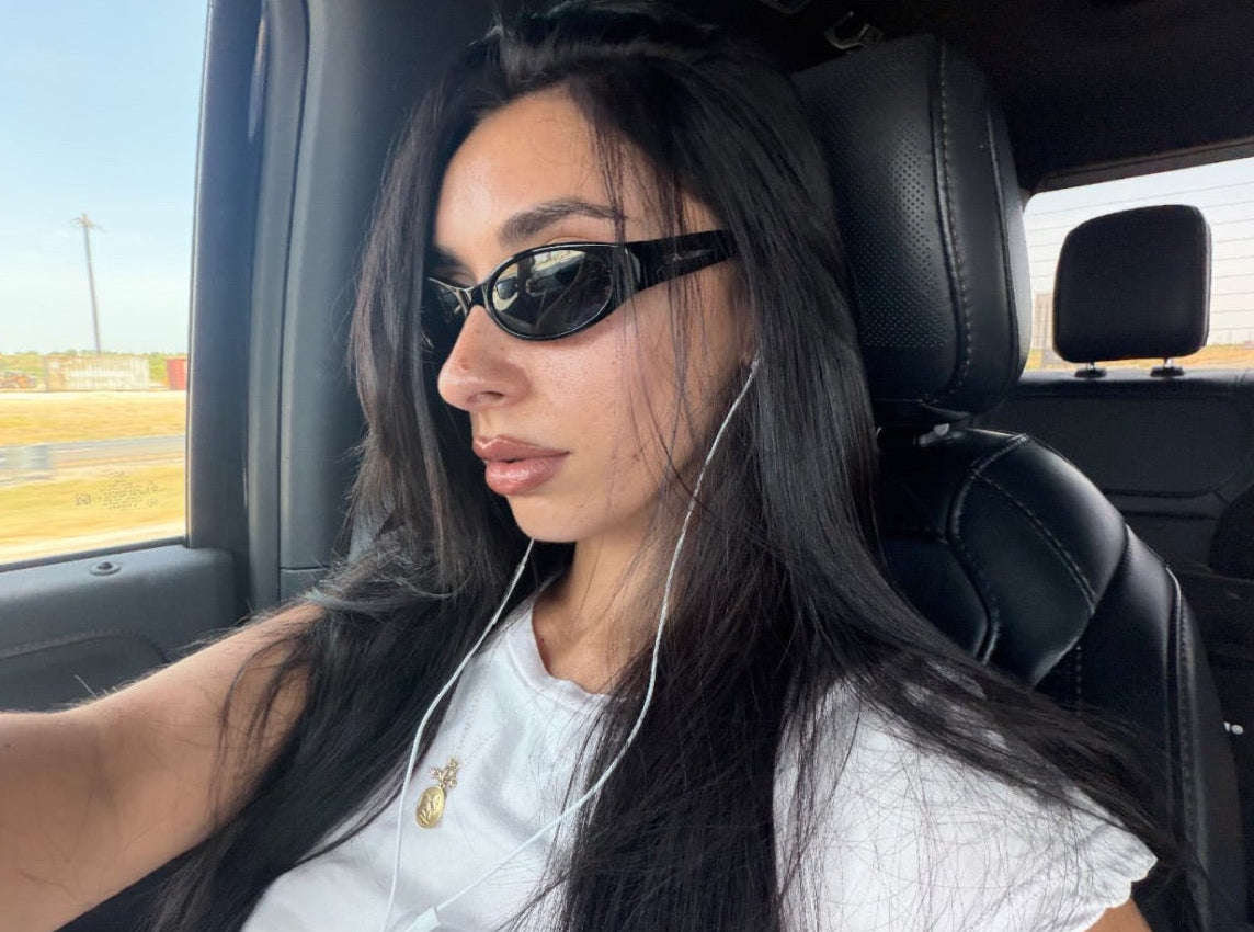 A social media post screenshot showing a woman with long black hair wearing black DAISY sunglasses and a white T-shirt inside a car.