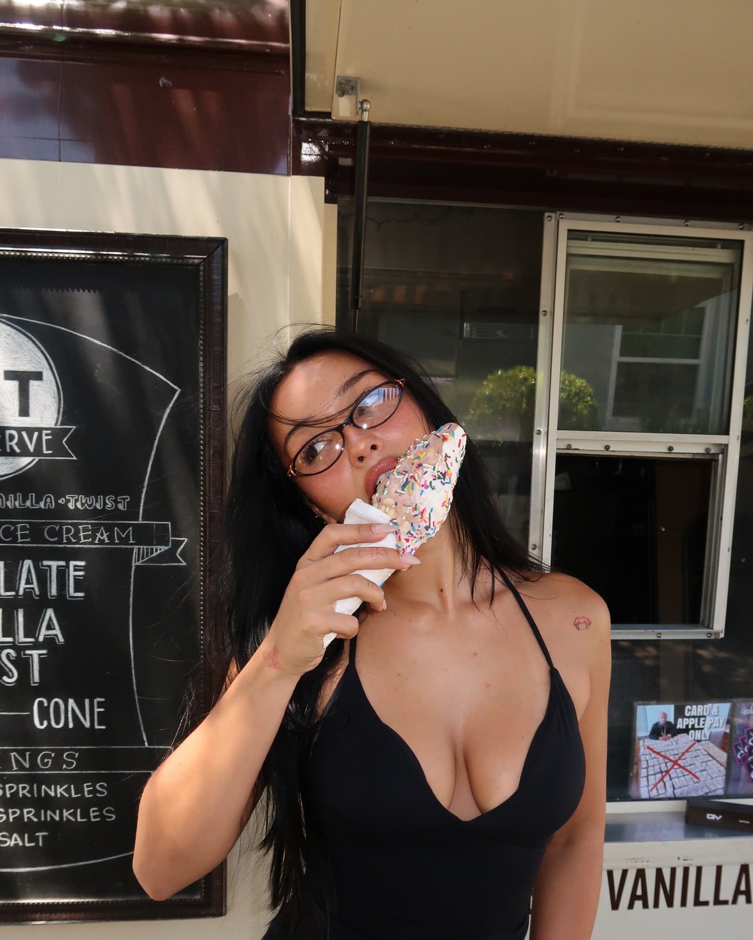 A woman with BANDAGE glasses and long dark hair wearing a black halter top and patterned shorts, holding a chocolate chip cookie in front of her face while standing in front of a vanilla ice cream truck.