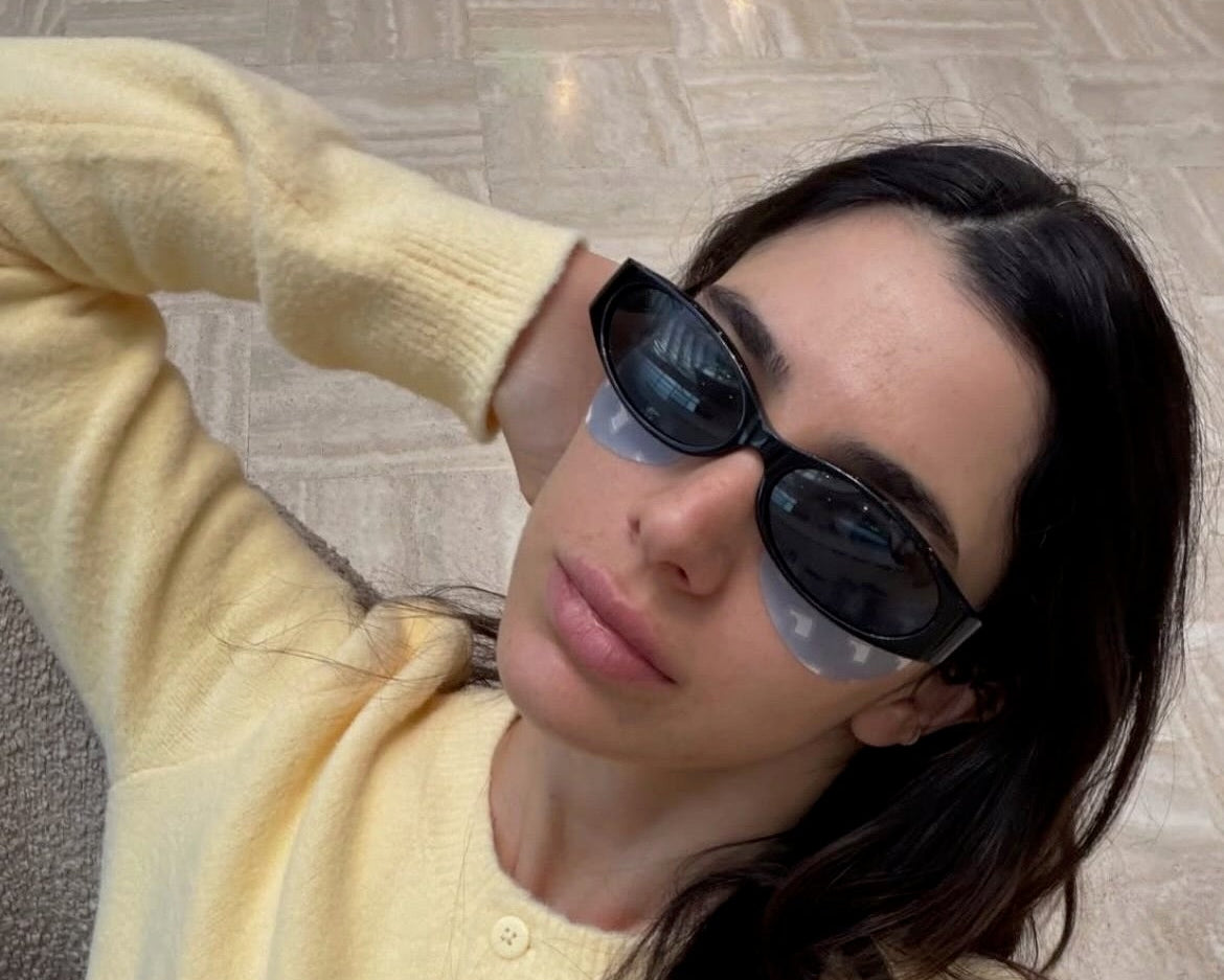 A woman wearing a pale yellow knit cardigan and black DAISY sunglasses takes a mirror selfie in an Instagram post. The cardigan has a button-down front and ribbed trim.