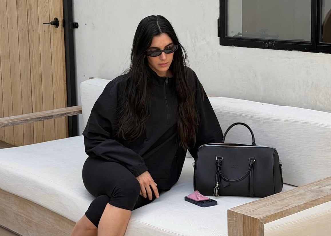 A woman wearing a black oversized sweatshirt and black capri leggings sits on a white outdoor daybed with a wooden frame. A black leather handbag rests beside her on the cushion.