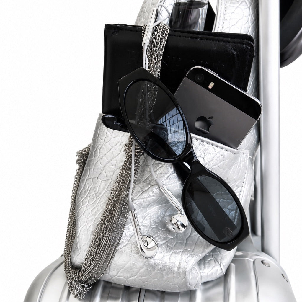 A silver textured shoulder bag with silver chain straps and silver-tone hardware, shown packed into a silver hard-shell suitcase. The bag contains black accessories, including a black wallet, a black smartphone, and black sunglasses with dark lenses.
