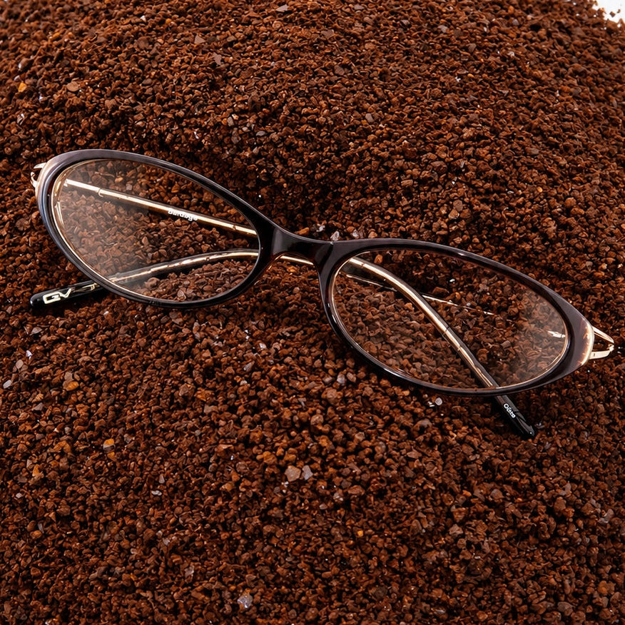 A pair of black cat-eye eyeglasses resting on a mound of dark brown coffee grounds against a plain white background.