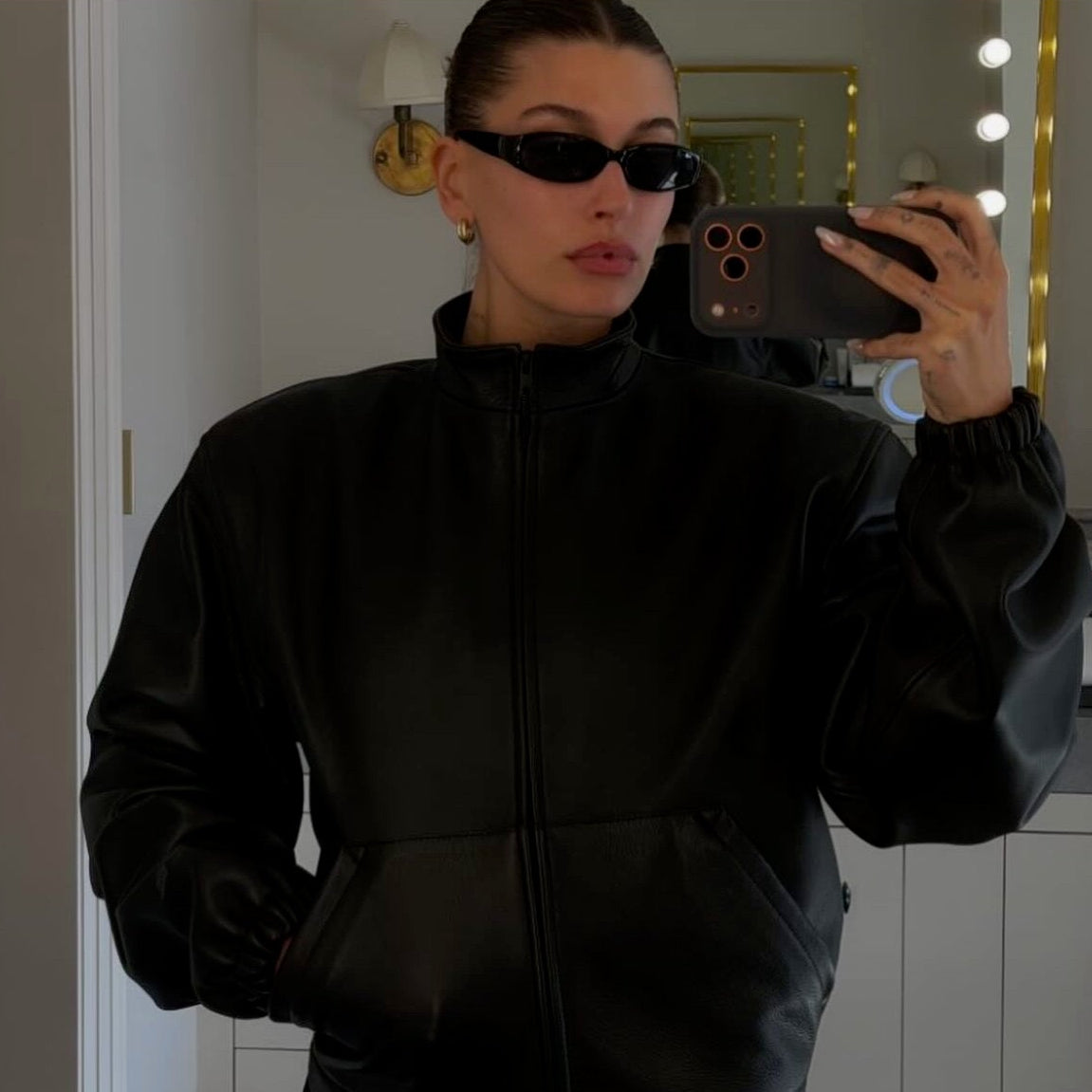 Hailey Beiber taking a mirror selfie wearing a black leather-look zip-up jacket and DAISY sunglasses, with an Instagram post interface visible around the photo.