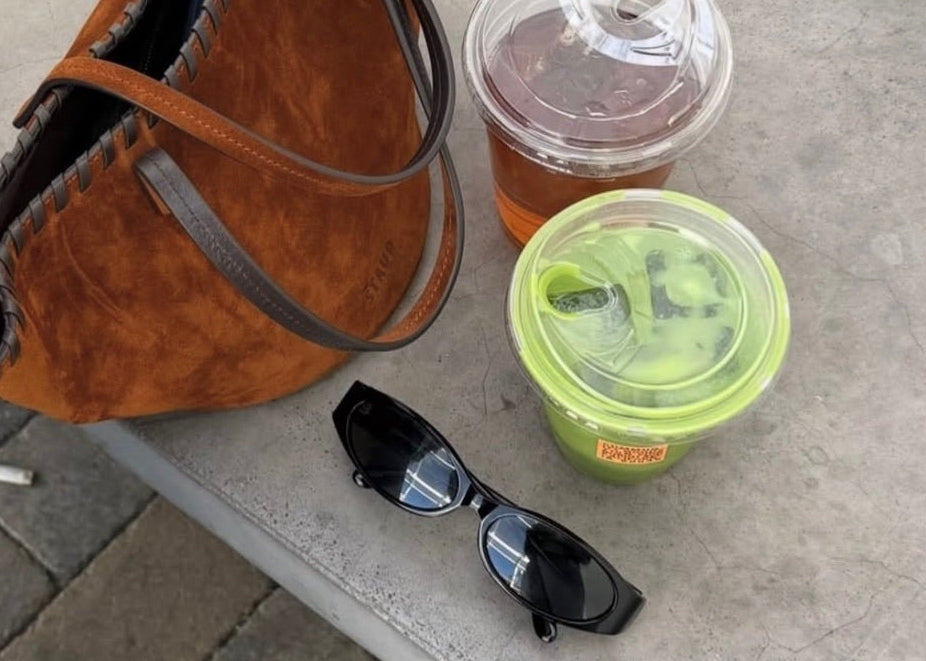 A brown suede tote bag with dark brown leather handles sits on a concrete bench next to sunglasses, a iced coffee, and a green-lidded drink container. A person wearing blue flats and light wash jeans is standing nearby on a paved sidewalk.