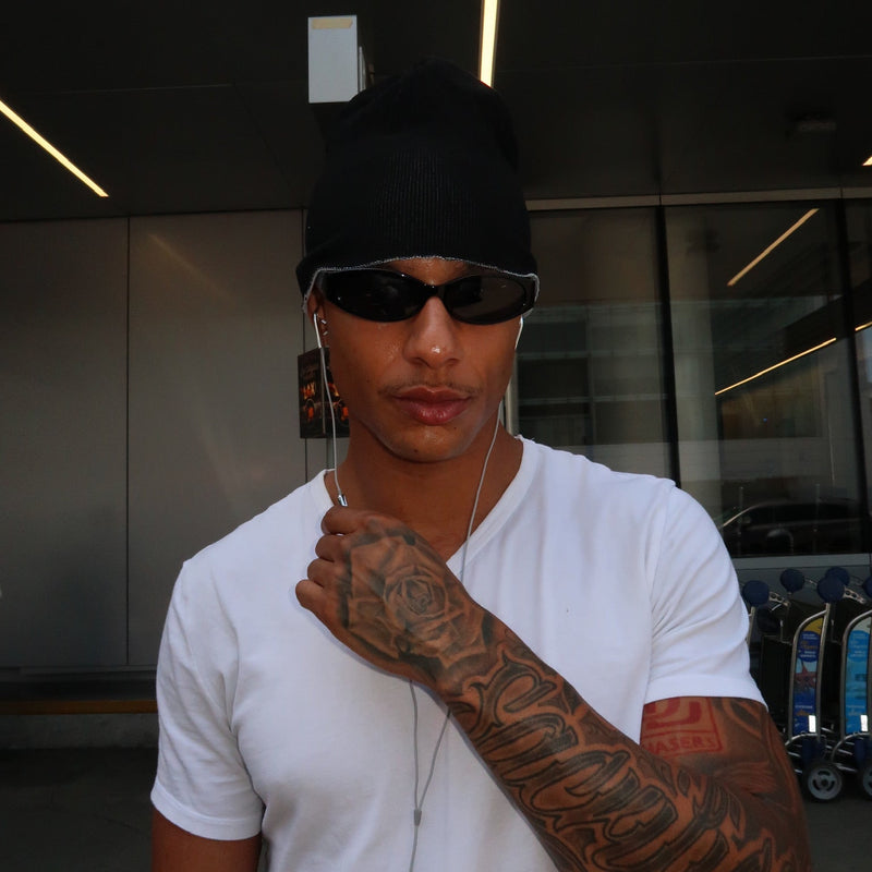 A man wearing a black beanie, black sunglasses, a white t-shirt, and white wired earphones, standing in front of a modern building entrance.