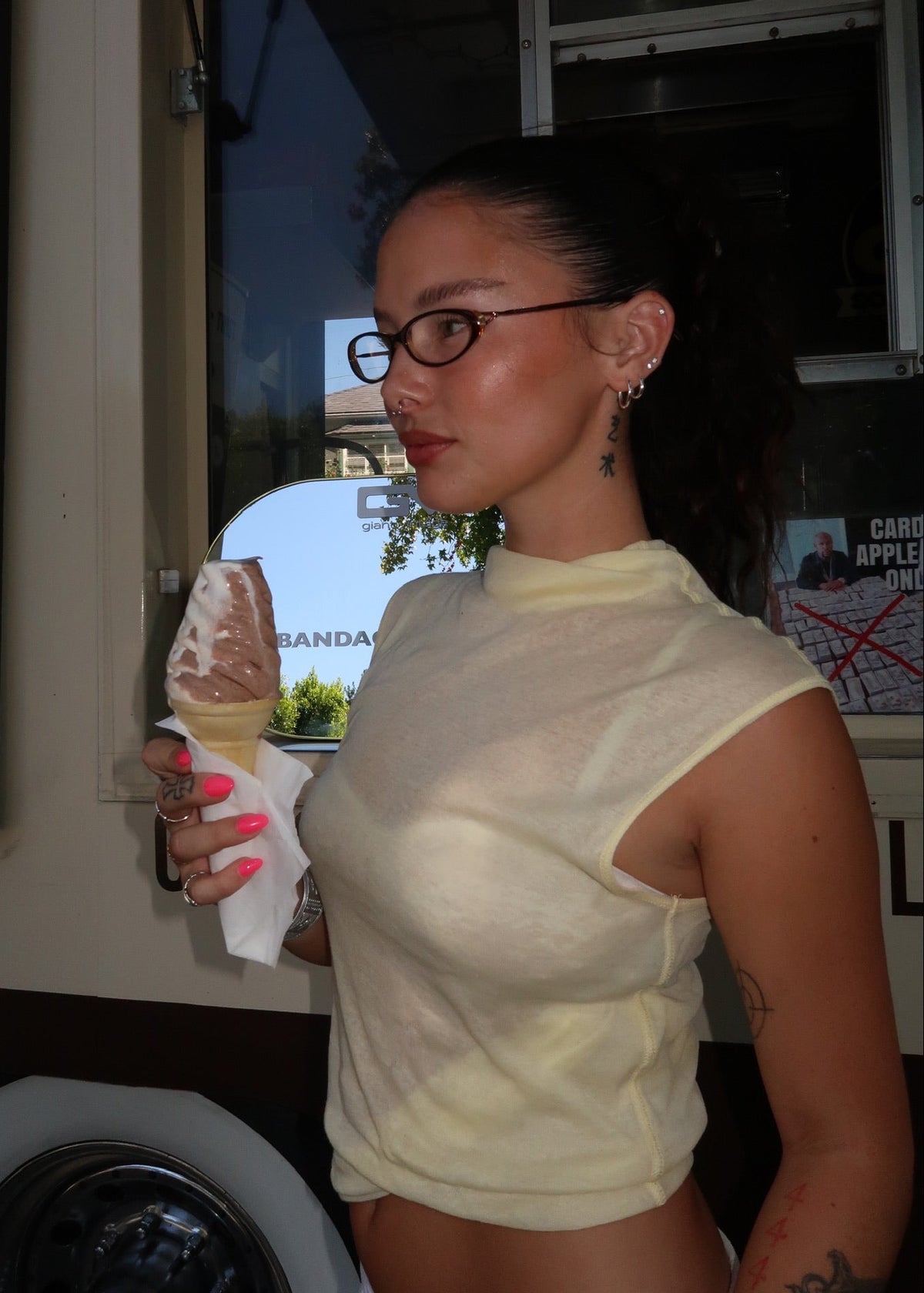 A woman with BANDAGE glasses and a ponytail stands indoors holding an ice cream cone. She is wearing a pale yellow sleeveless mock-neck crop top and white bottoms.
