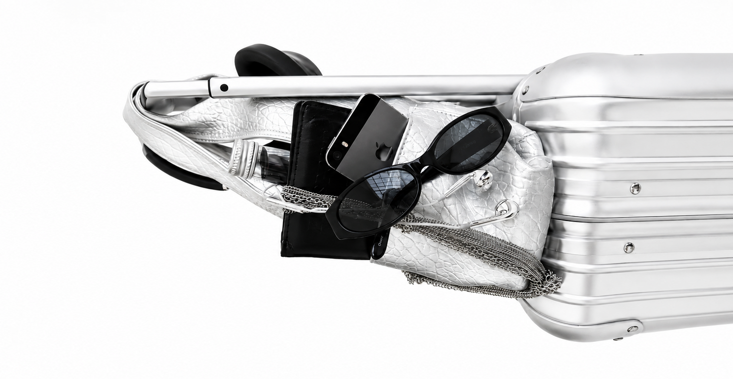 A silver metallic shoulder bag with a chain strap, shown packed onto the side of a matching silver hard-shell suitcase against a plain white background. The bag is styled with black accessories including sunglasses, a wallet, and a phone.
