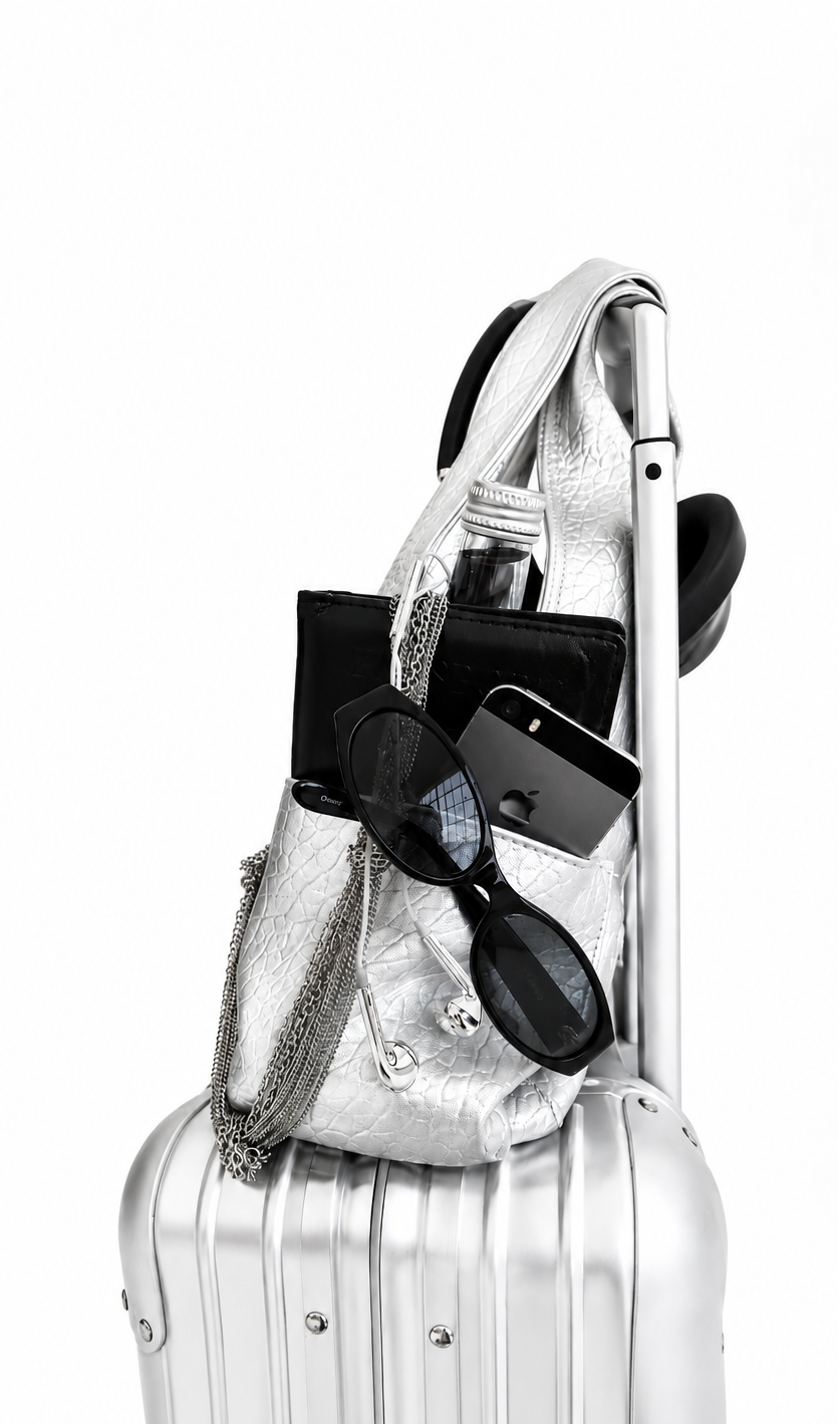 A silver metallic shoulder bag with a chain strap, shown packed onto the side of a matching silver hard-shell suitcase against a plain white background. The bag is styled with black accessories including sunglasses, a wallet, and a phone.