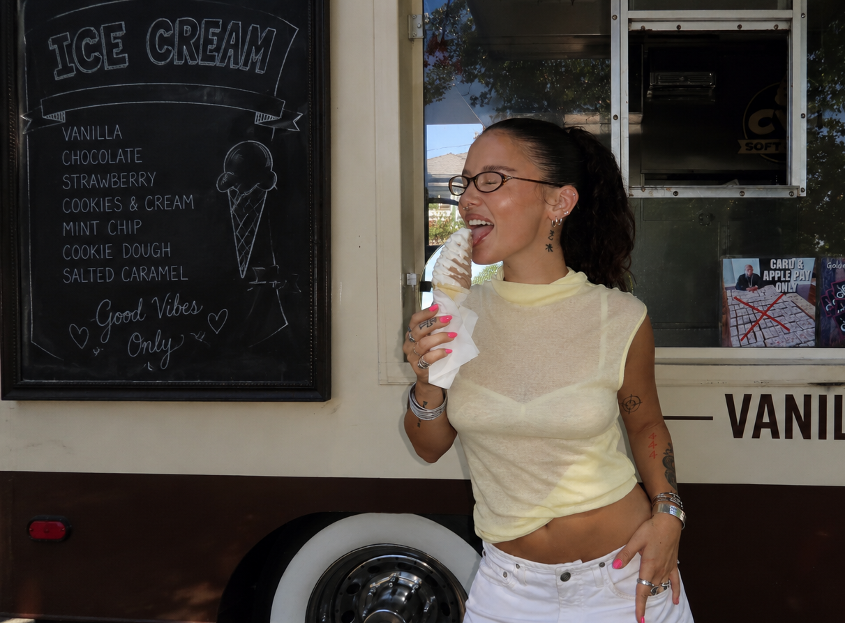 A woman wearing BANDAGE glasses, a sheer pale yellow top, and white shorts stands in front of a vanilla ice cream truck, holding a cone of ice cream and smiling. The truck has a black chalkboard menu listing flavors like vanilla, chocolate, strawberry, and cookie dough.