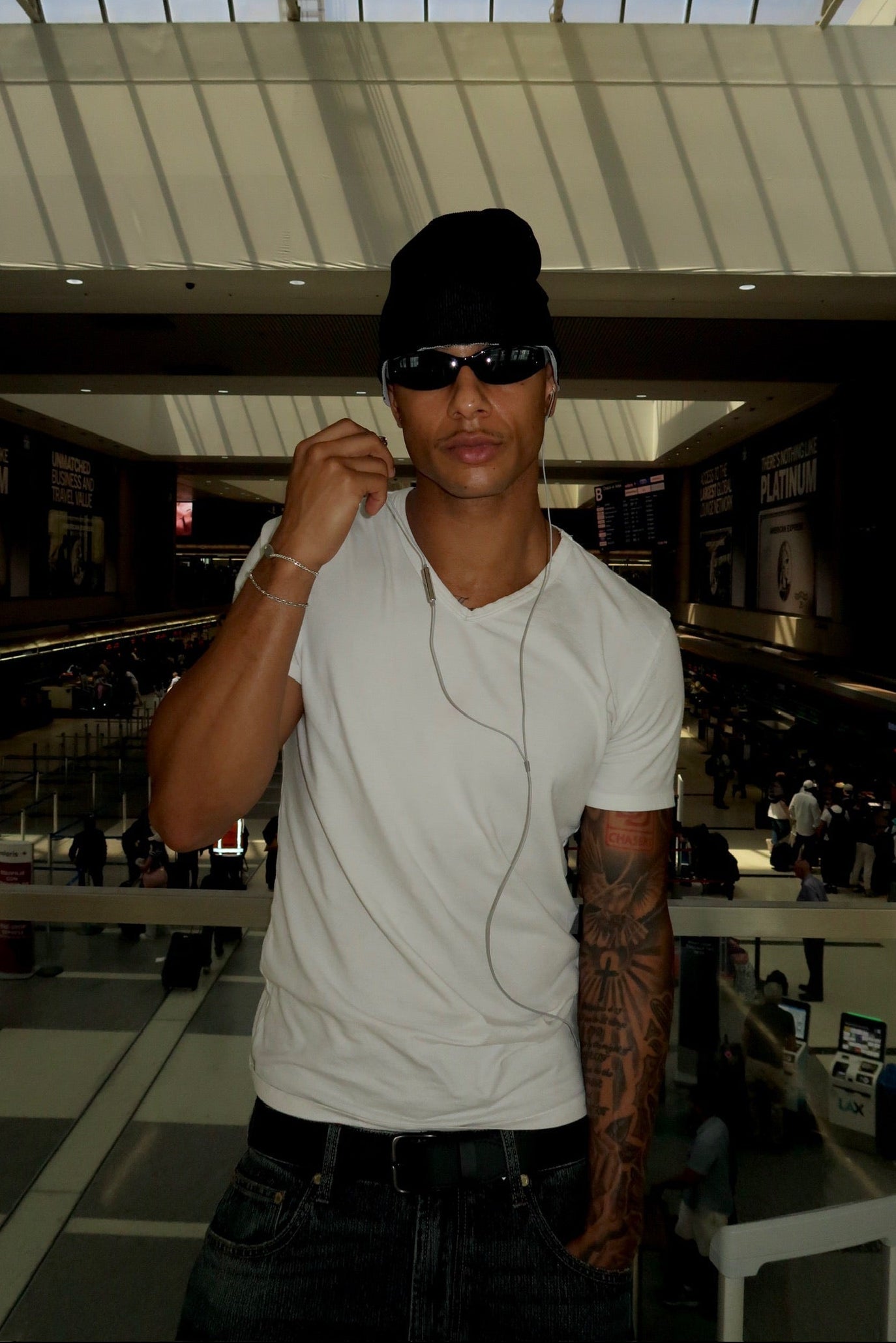 A man wearing a white t-shirt, black beanie, black sunglasses, and dark jeans stands in a modern indoor space with a glass roof and atrium.