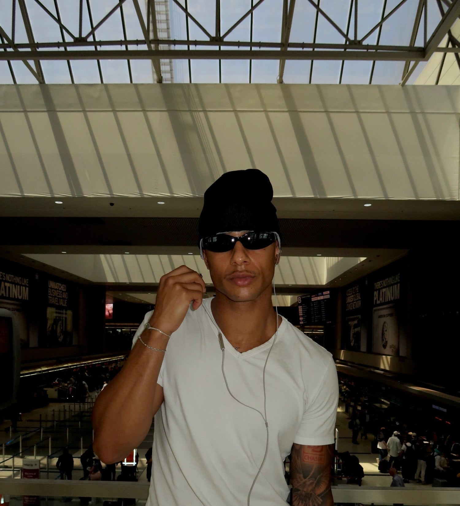 A man wearing a white t-shirt, black beanie, black sunglasses, and dark jeans stands in a modern indoor space with a glass roof and atrium.