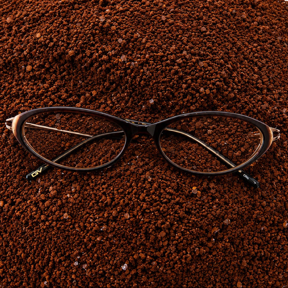 Black cat-eye eyeglasses resting on a large mound of dark brown coffee grounds against a white background.