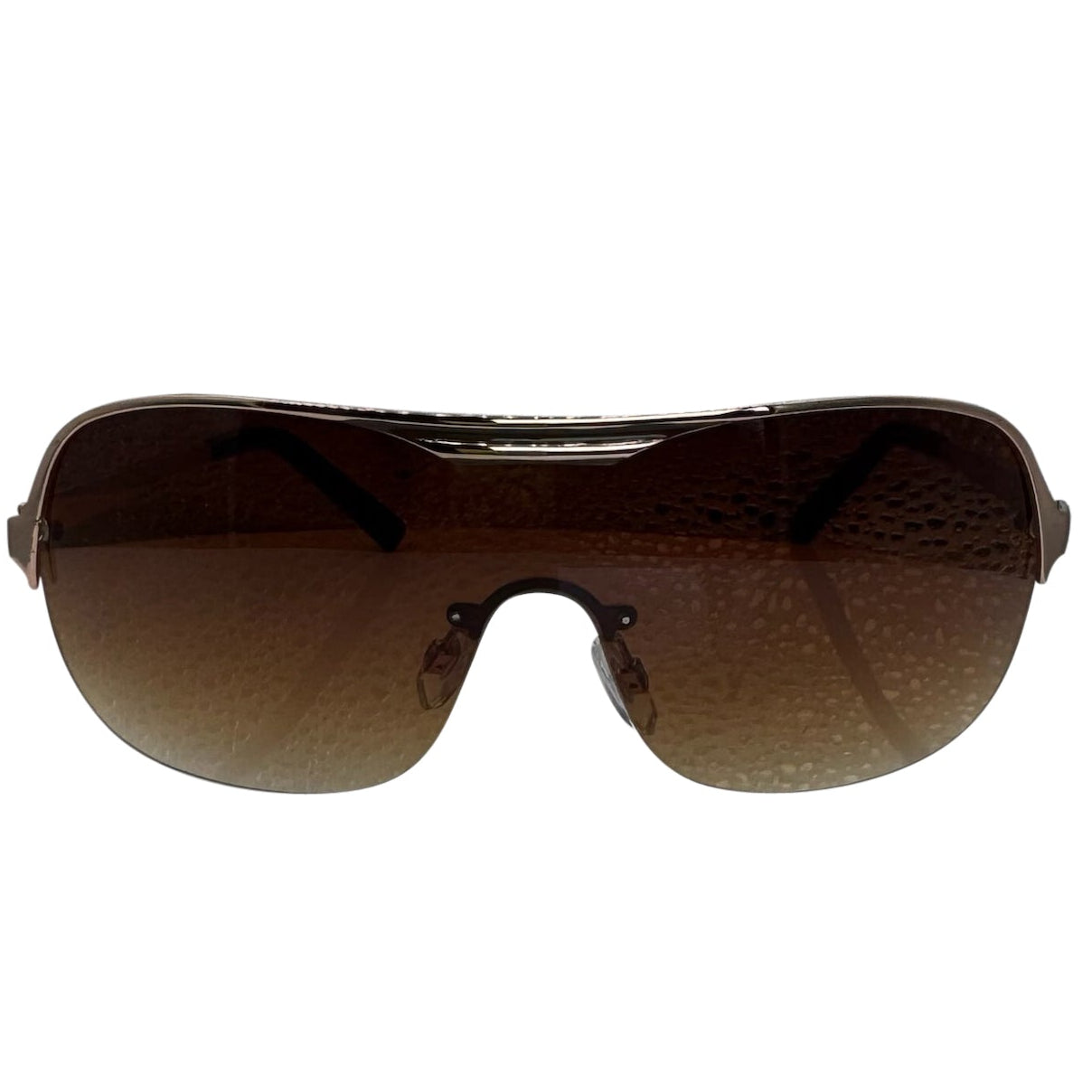 A front-facing product shot of the EXOTIC sunglasses, featuring a rimless shield design with oversized, dark-to-light gradient lenses. The frames are accented by a sleek brown/ amber double-bridge bar across the top and white arms visible through the lenses.