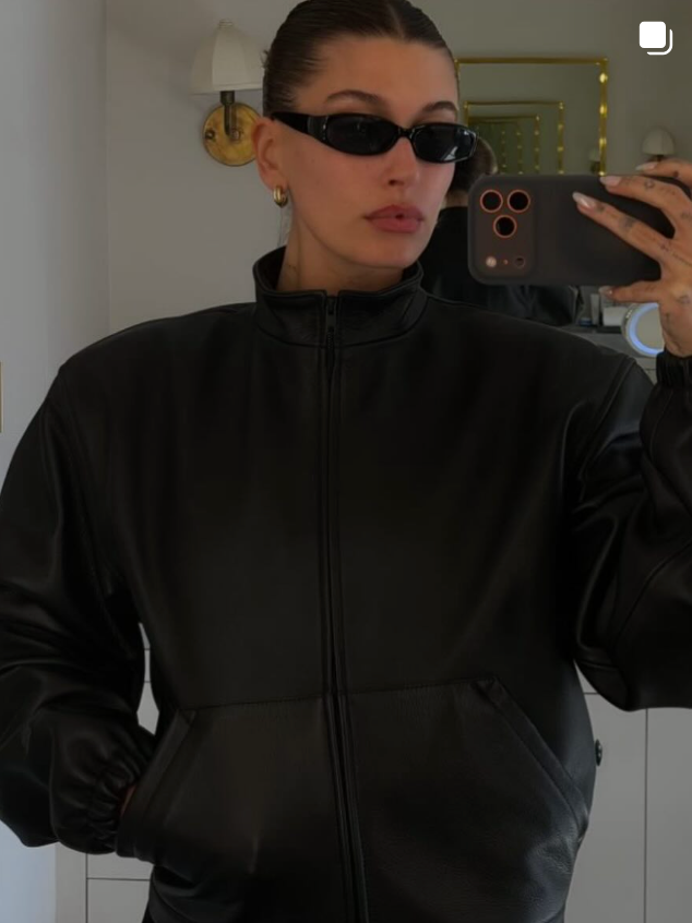 hailey beiber wearing a black jacket and sunglasses taking a mirror selfie.