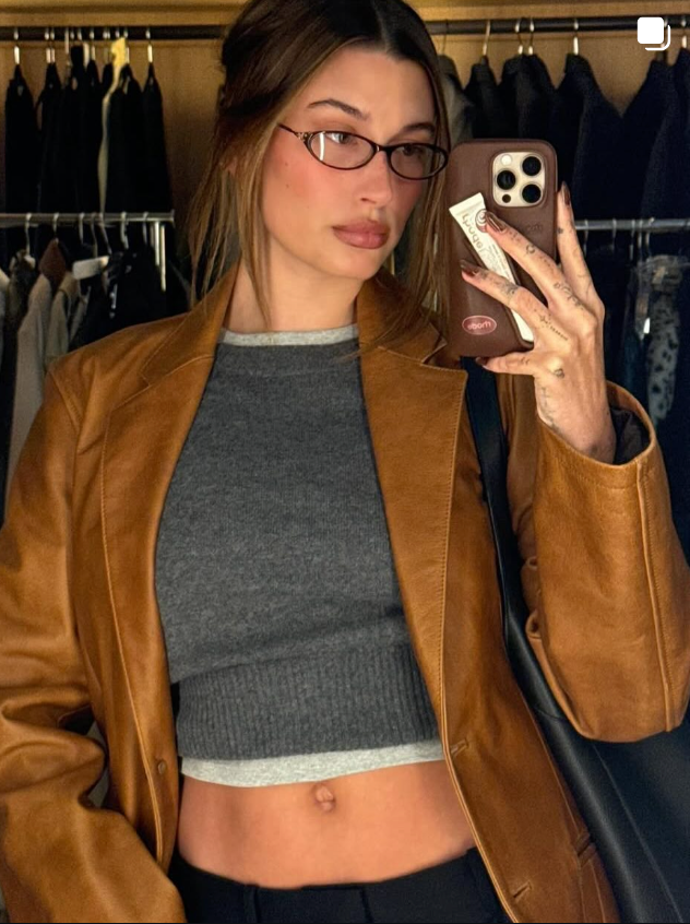 Hailey bieber wearing a brown leather jacket and gray top, taking a mirror selfie in a clothing store.