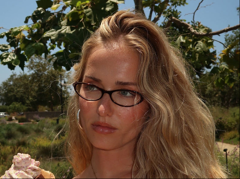 A woman with long blonde hair and glasses, wearing a white tank top, holds an ice cream cone outdoors under a blue sky with trees in the background.