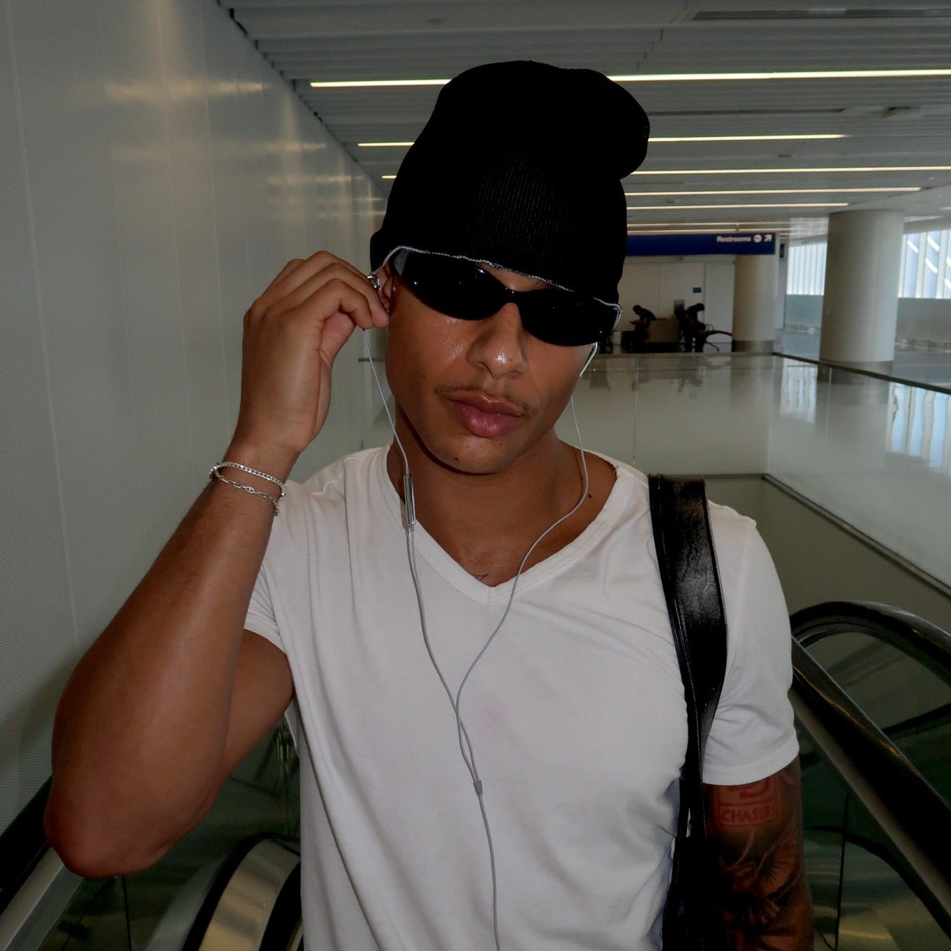 A man wearing a black beanie, black sunglasses, and a white t-shirt standing on an escalator in a modern indoor hallway.