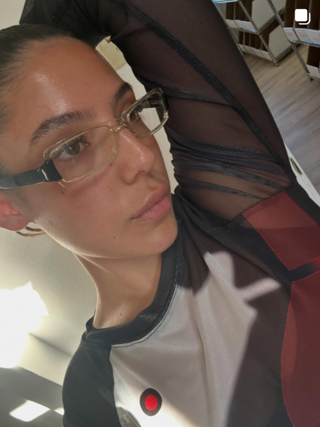 Person wearing glasses and a dark outfit with a red button, indoors.