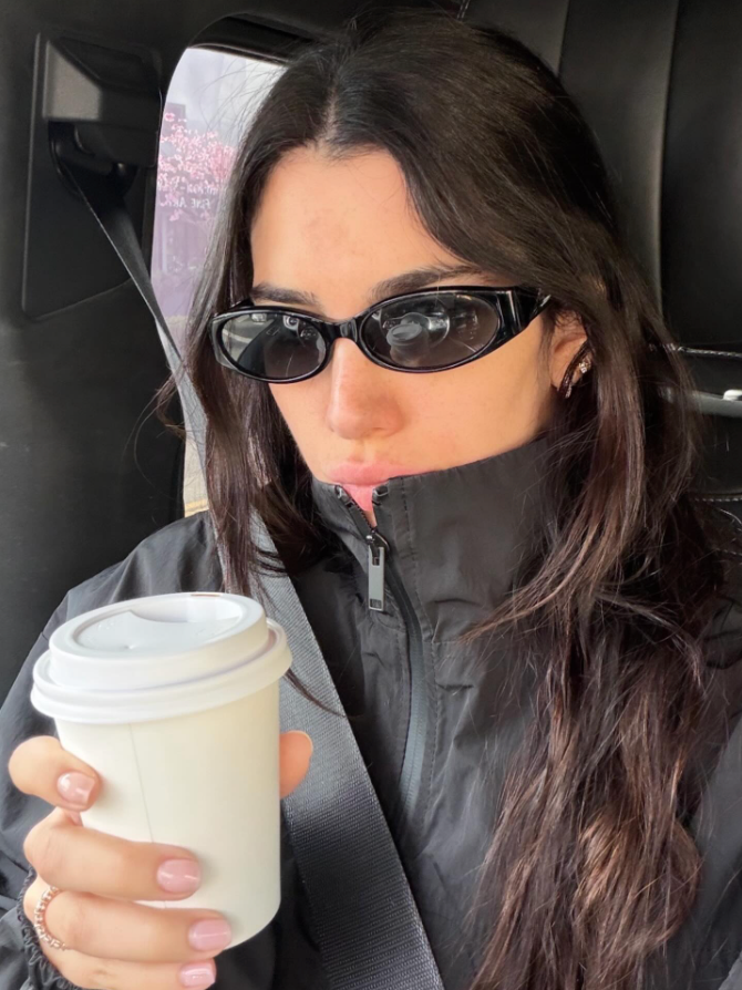 Woman in a car holding a coffee cup, wearing sunglasses and a dark jacket.