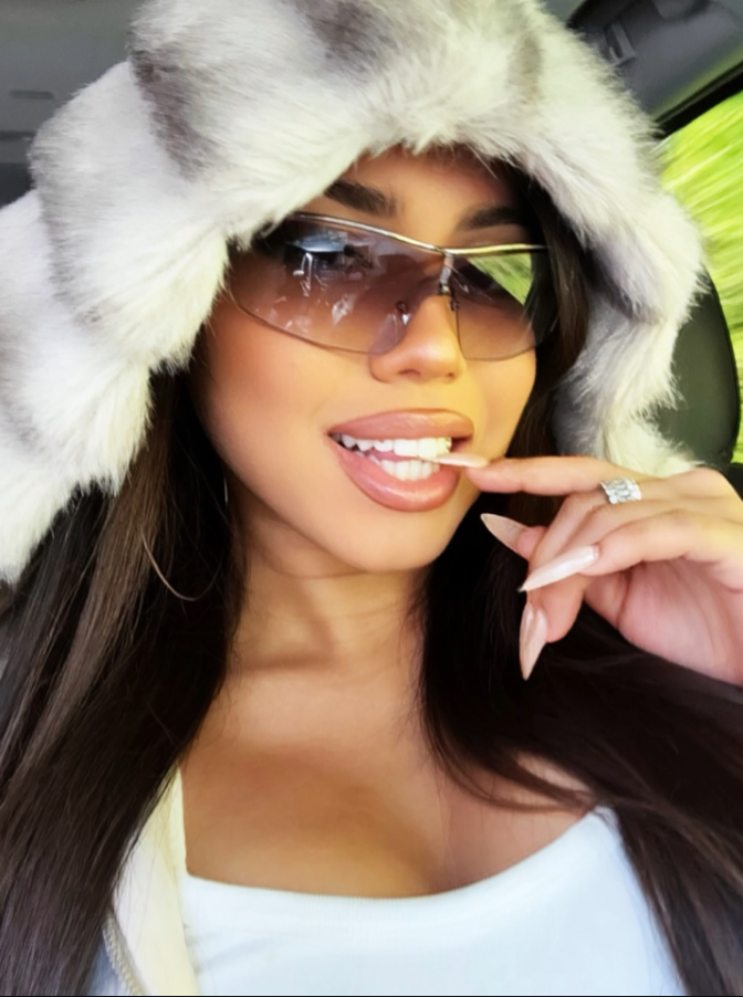 Woman wearing a fur-lined hood and sunglasses, posing with a hand gesture.