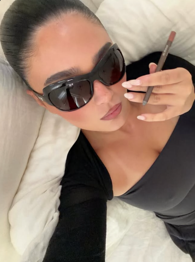 Person wearing sunglasses and holding a makeup brush, sitting on a white couch.