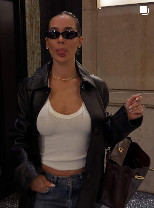Person wearing a black leather jacket, white crop top, and blue jeans, holding a brown bag.